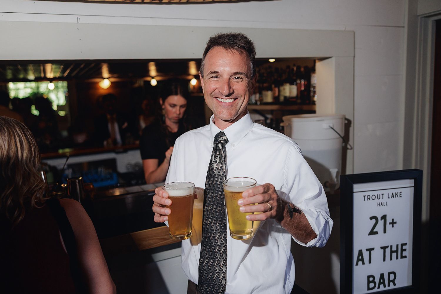 A person in business attire holding two pint glasses of beer at a bar with a 21+ sign visible in the background.