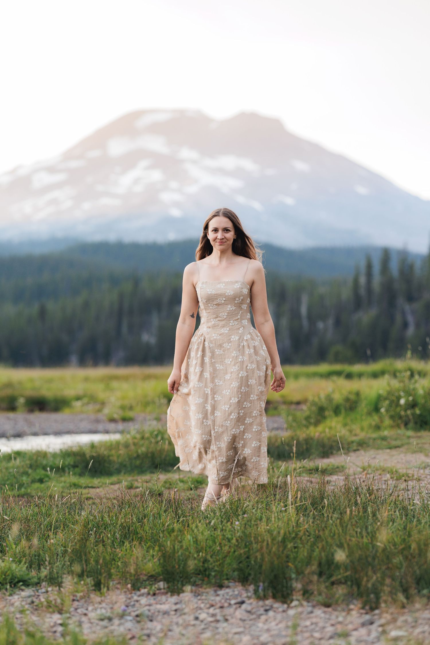 A person in a cream-colored dress walks through a meadow against a backdrop of majestic mountain peaks.