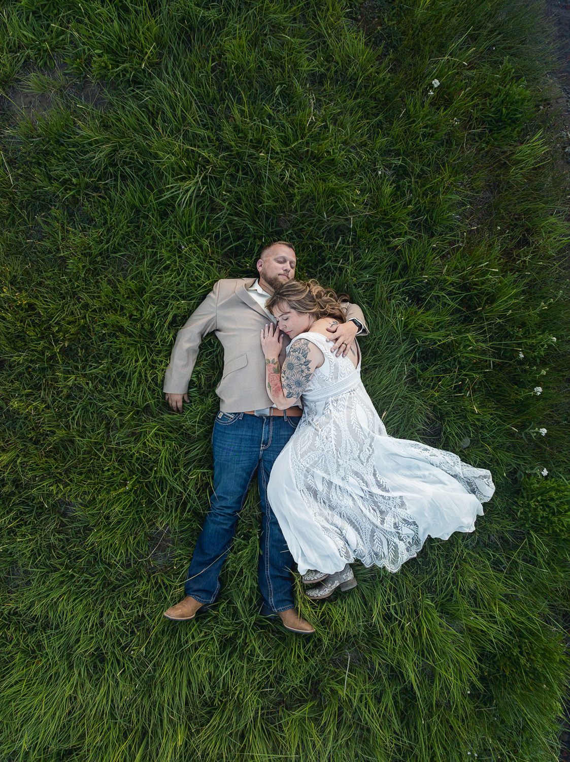 A couple in wedding attire lies peacefully in lush green grass during an outdoor photoshoot.
