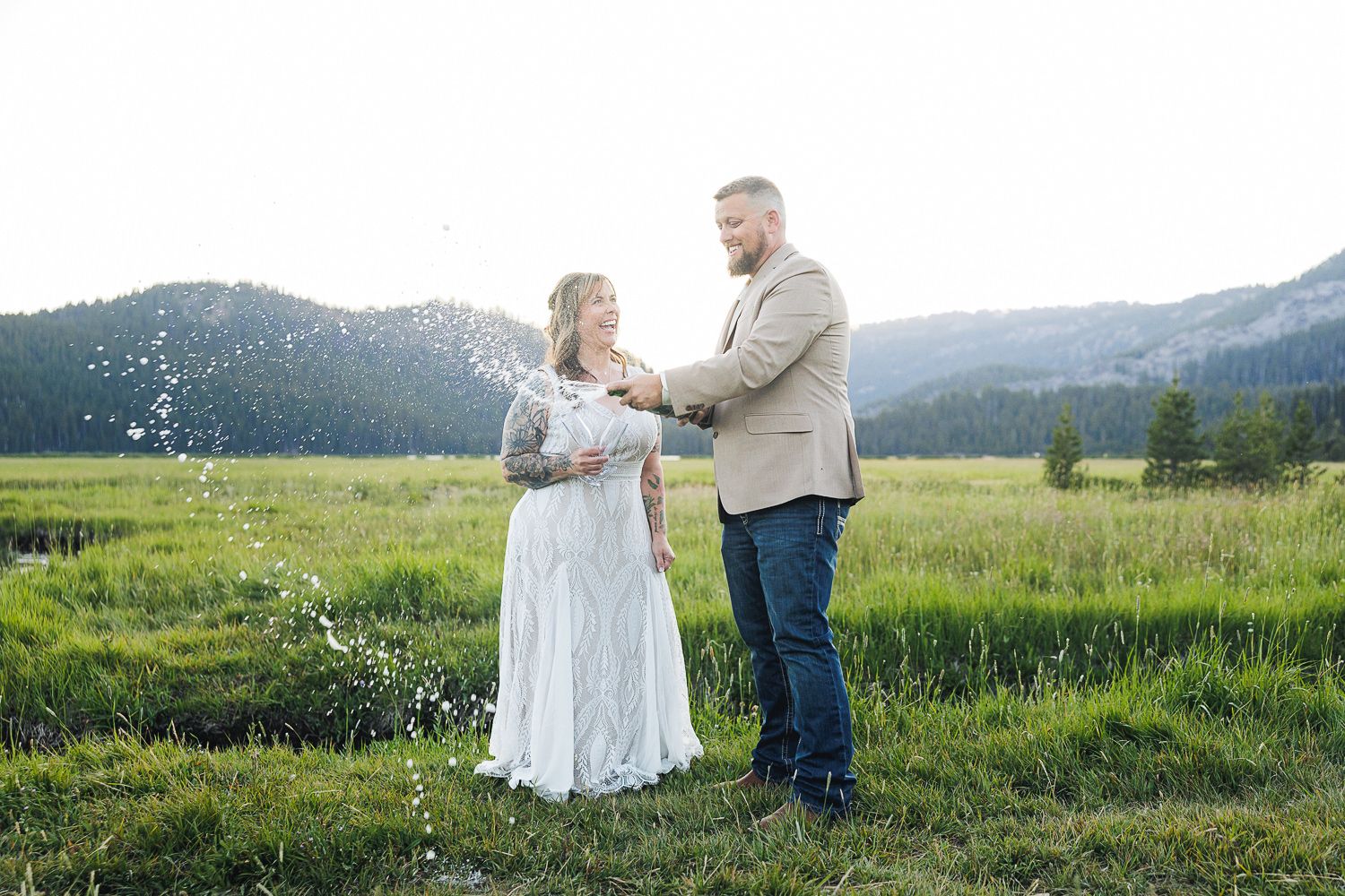 A couple stands together in a scenic mountain meadow with wildflowers at sunset in a romantic embrace.