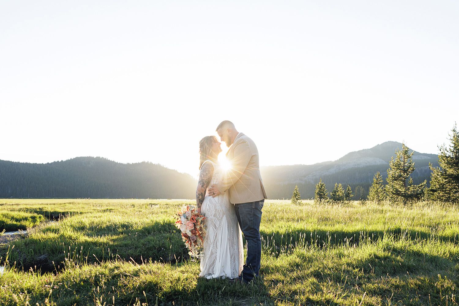 Two people embrace in a romantic moment at sunset in a mountain meadow with sunlight streaming through.