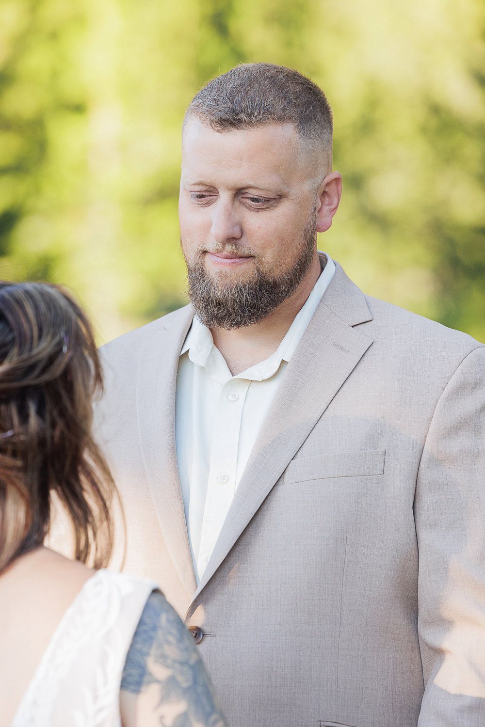 Groom wearing a light gray suit and white shirt looks down thoughtfully during an outdoor wedding ceremony.