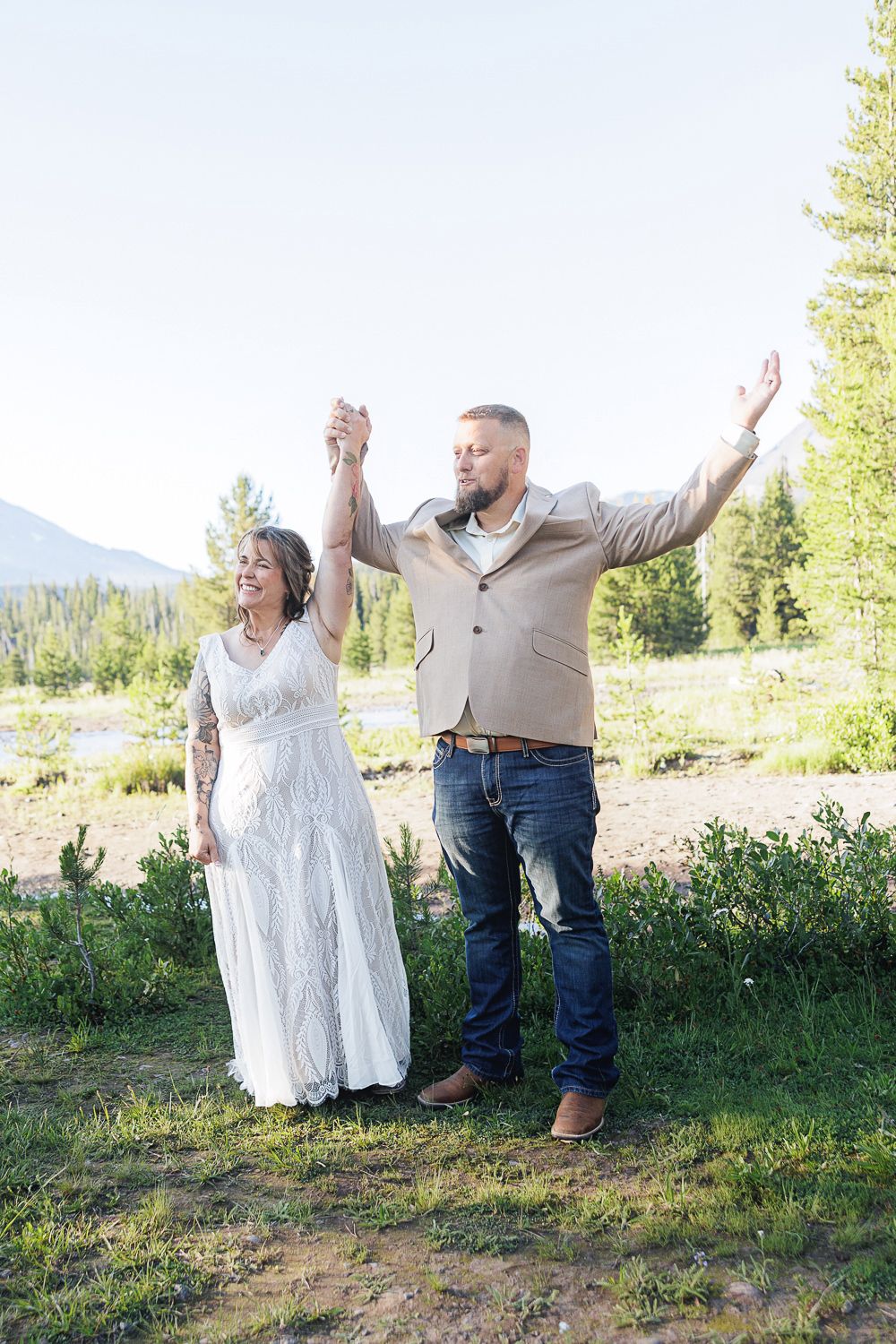 A joyful couple celebrates outdoors in a mountainous landscape with trees, the bride wearing a flowing white dress.