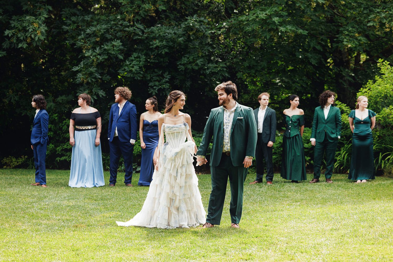 Bride and groom hold hands in a lush garden surrounded by wedding party in blue and green attire.