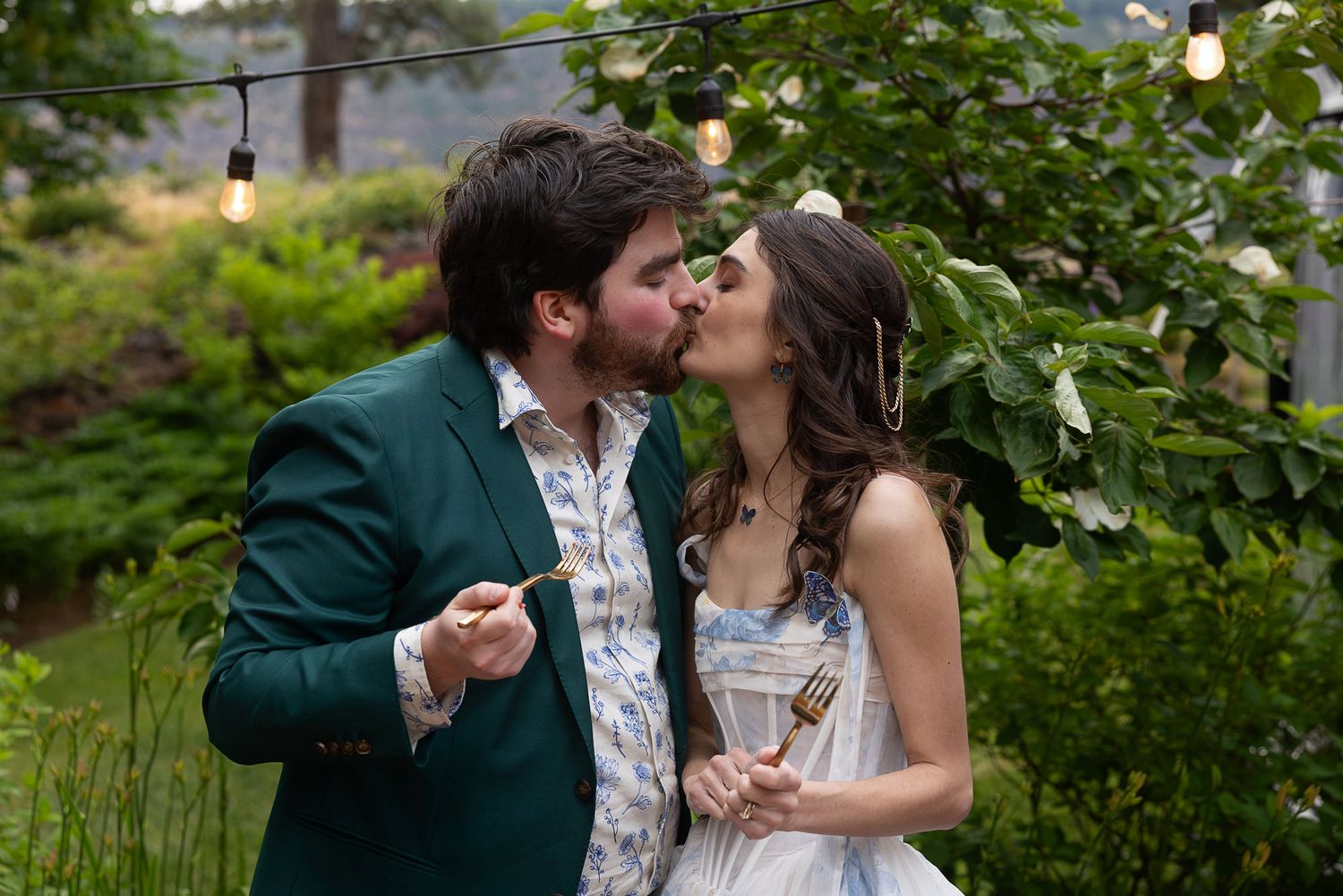Couple sharing a romantic kiss at an outdoor garden wedding, man in green suit, woman in blue floral dress.