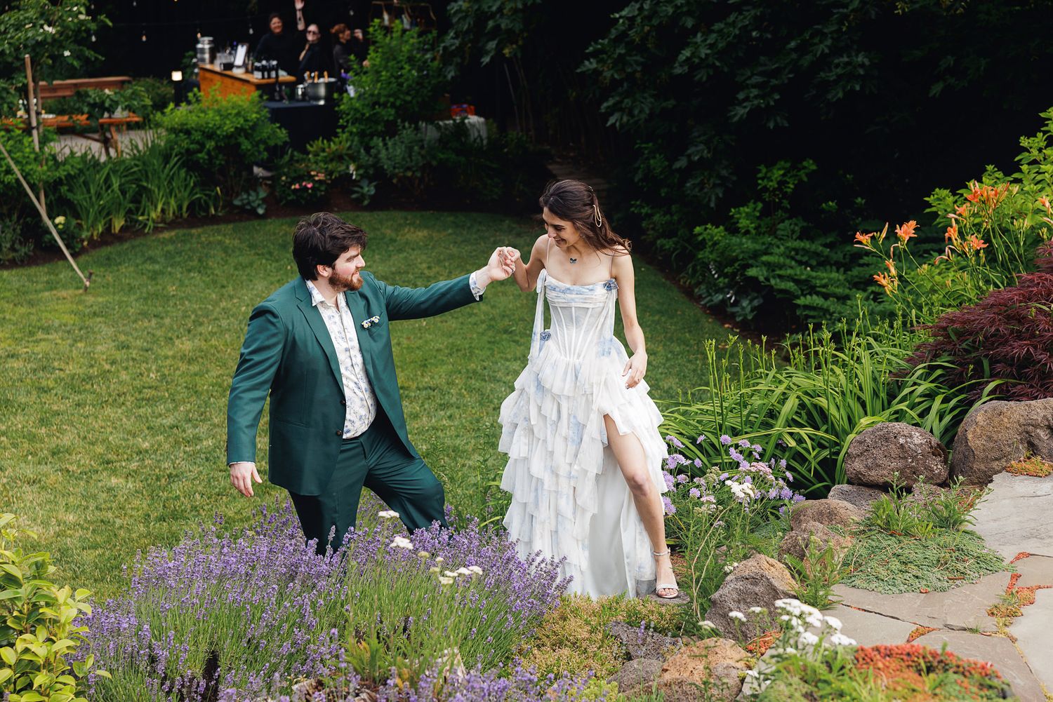 Groom in green suit kneeling to hold bride's hand in a lush garden wedding setting.
