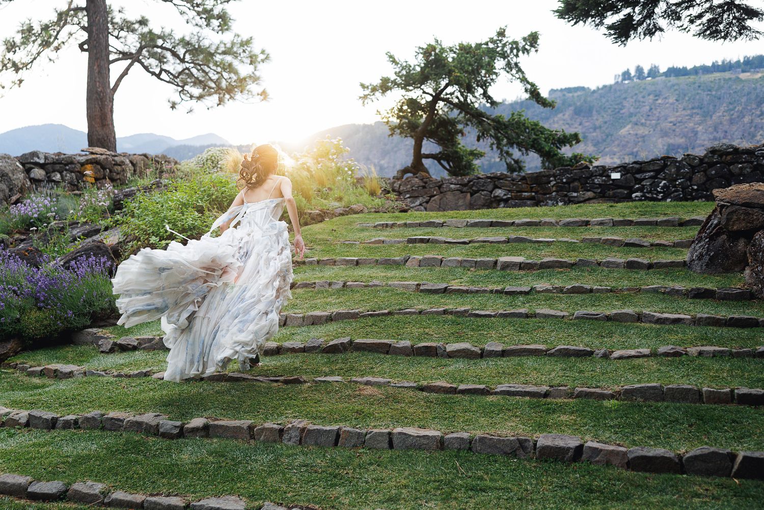 Bride in flowing white gown ascending stone steps at sunset with mountain views and lavender gardens.
