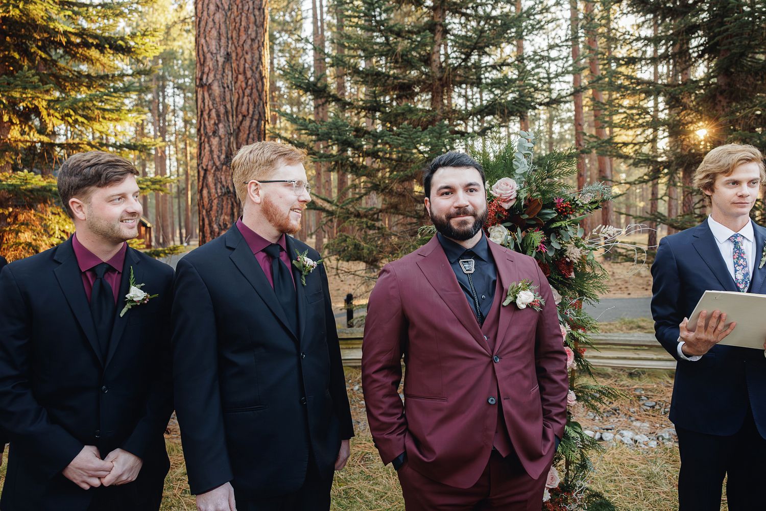 A group in formal attire stands together in a sunlit forest setting during an outdoor ceremony.