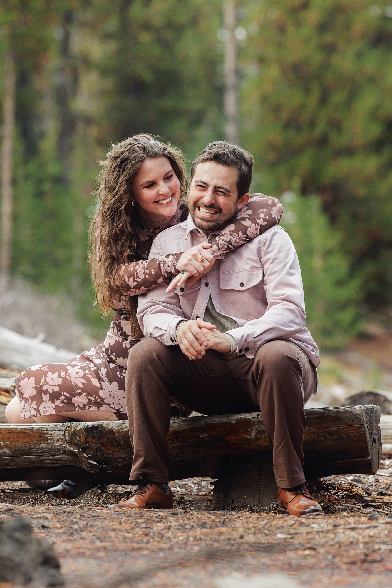 A couple relaxes and laughs together while sitting on a fallen log in an autumn forest setting.