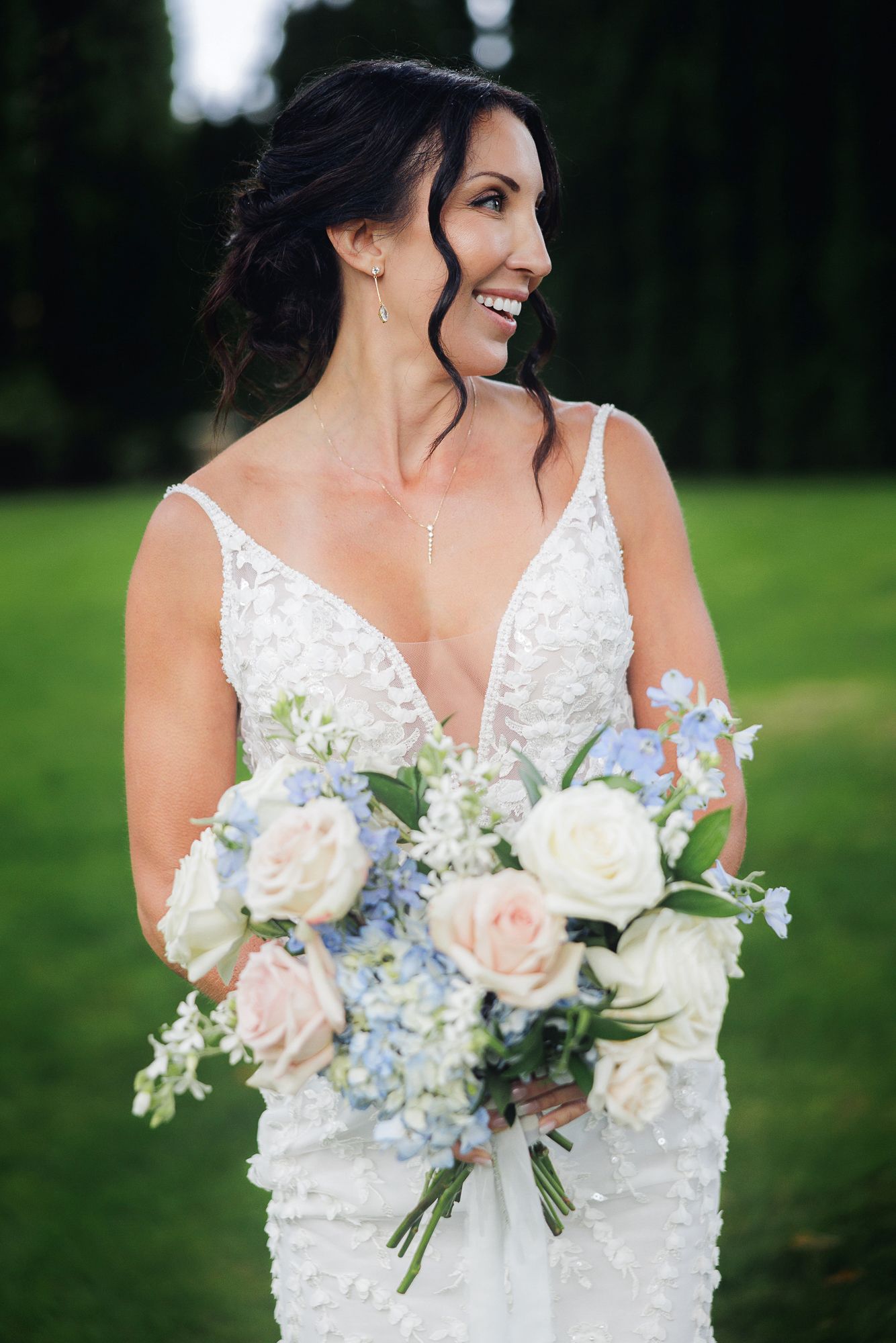 Bride in lace wedding dress holds elegant bouquet of white roses and blue hydrangeas against green outdoor backdrop.