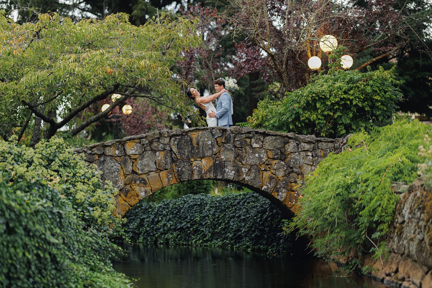 A romantic moment captured on a rustic stone bridge surrounded by lush greenery and hanging lanterns.