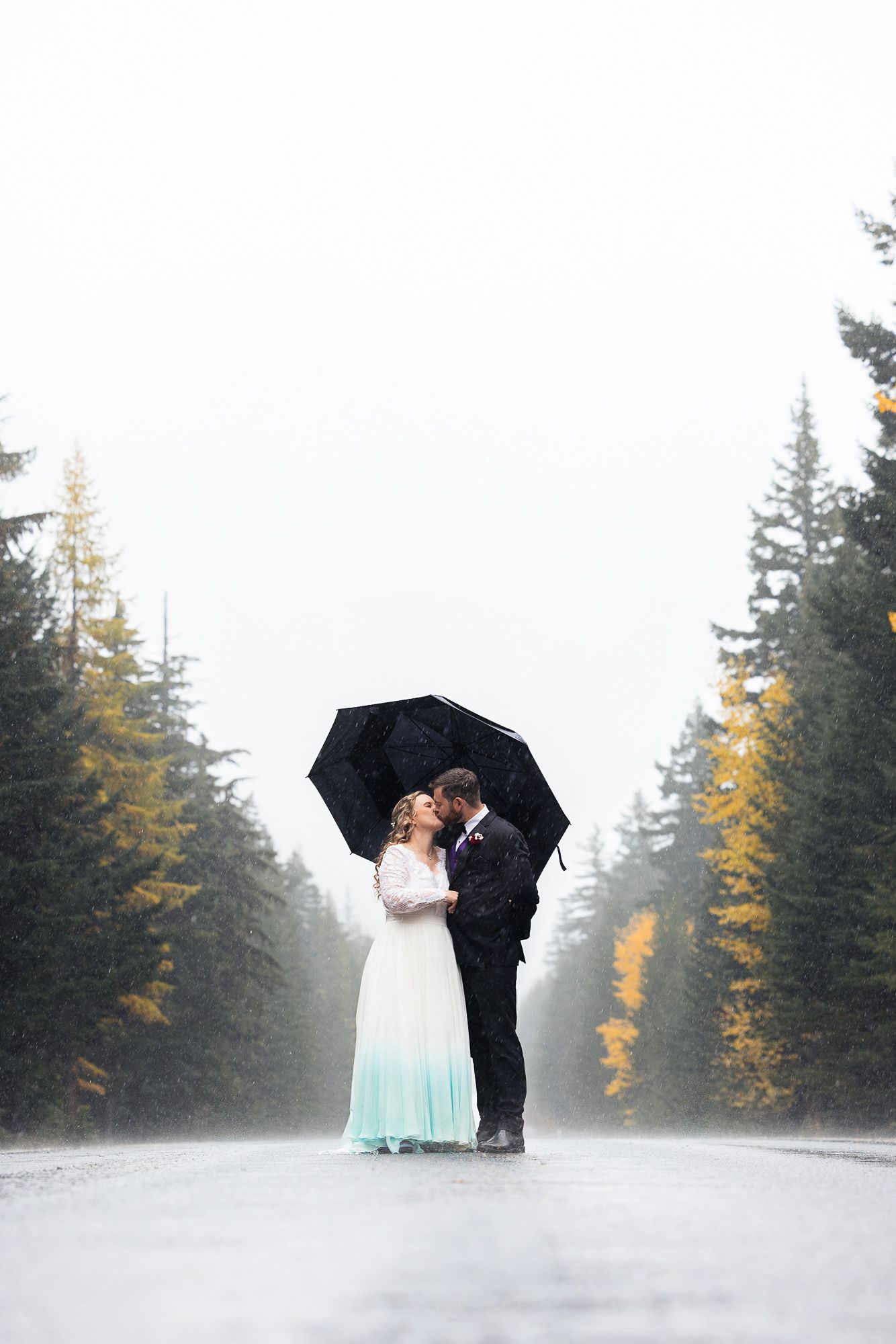 A couple shares a romantic moment under a black umbrella on a misty forest road with autumn trees in the background.