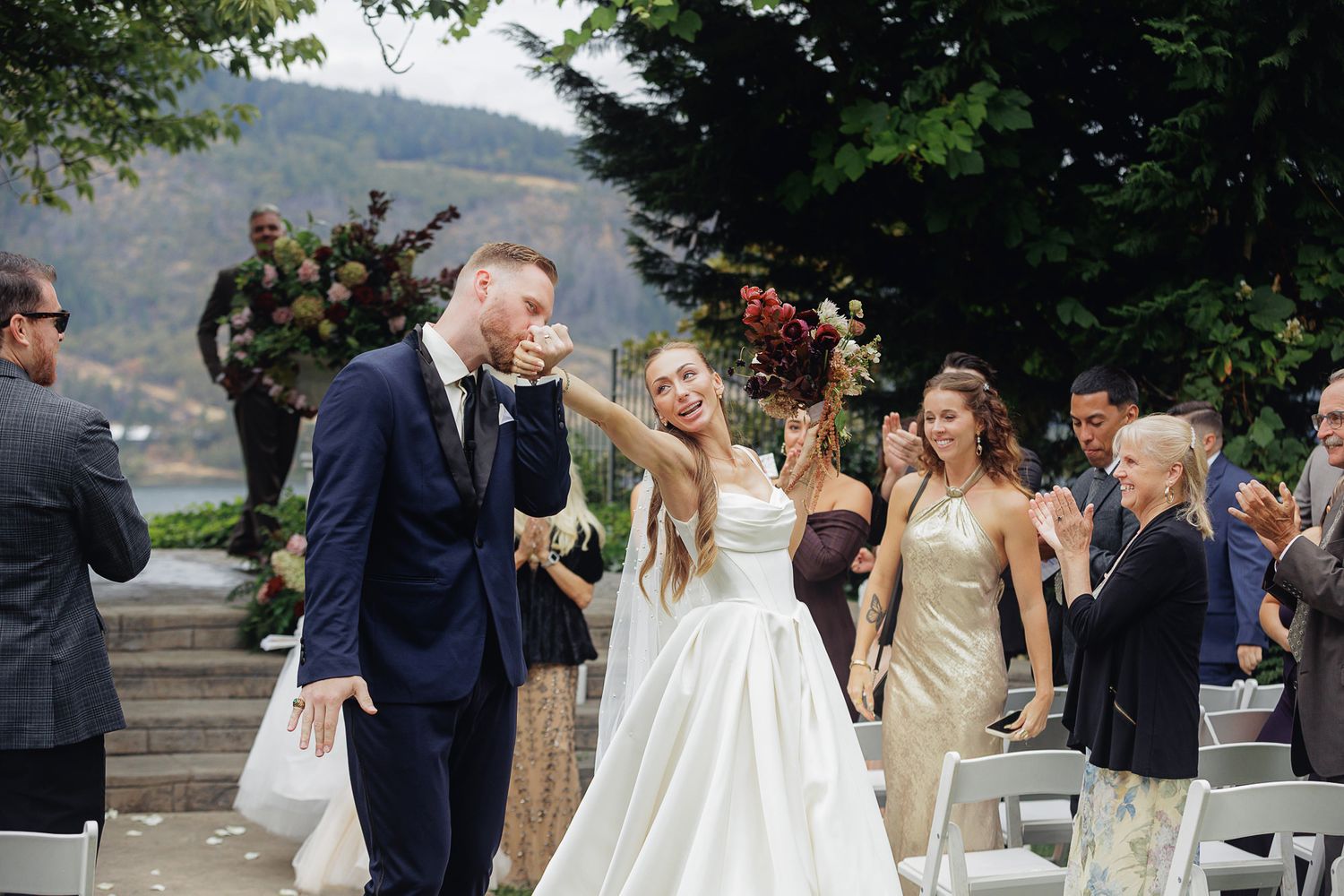 Bride and groom walk down the aisle after wedding ceremony outdoors with guests applauding.