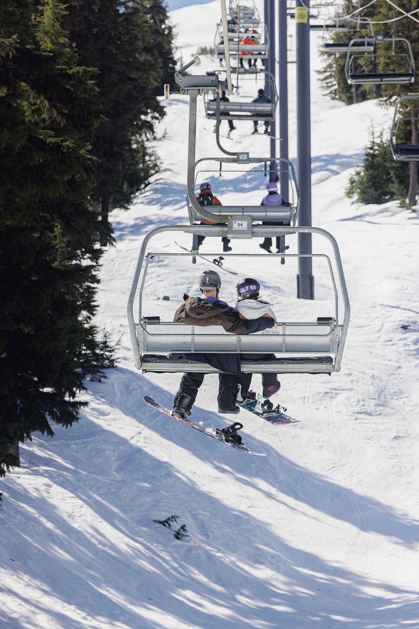 Two skiers ride up a ski lift on a snowy mountain slope with trees in the background.