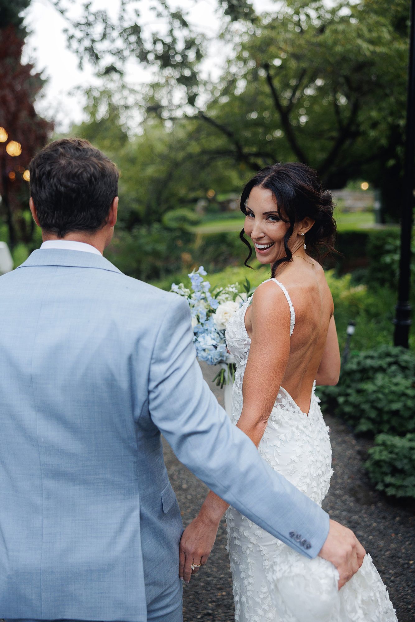 Newlywed couple walking and laughing together in a garden setting, bride in white dress and groom in light blue suit.