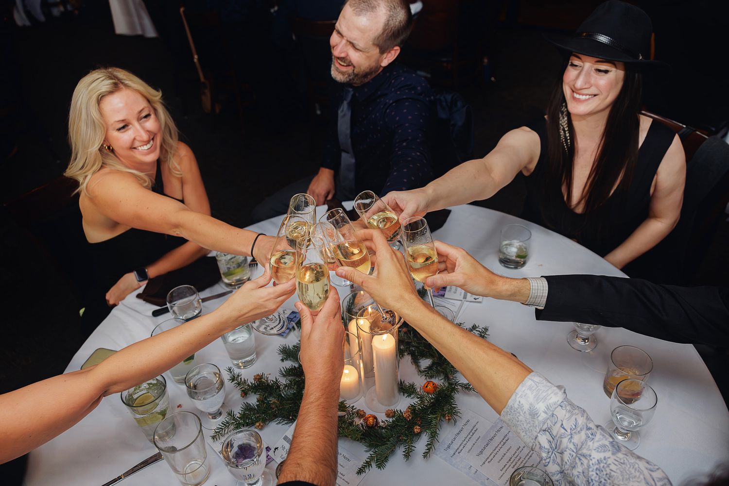 Wedding reception guests reach across a formal dining table to clink their wine glasses in celebration.