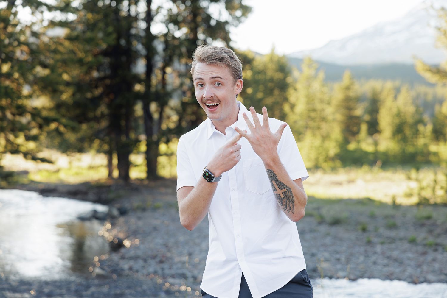 Young man in white shirt smiling outdoors near mountain stream, showing tattoos and smartwatch in scenic forest setting.