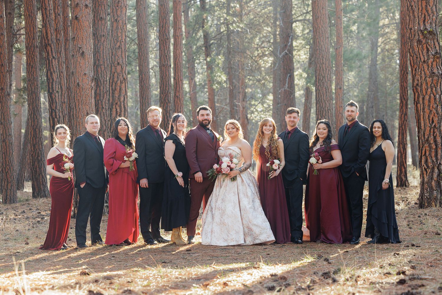 A wedding party poses in a line among tall pine trees in a forest setting.