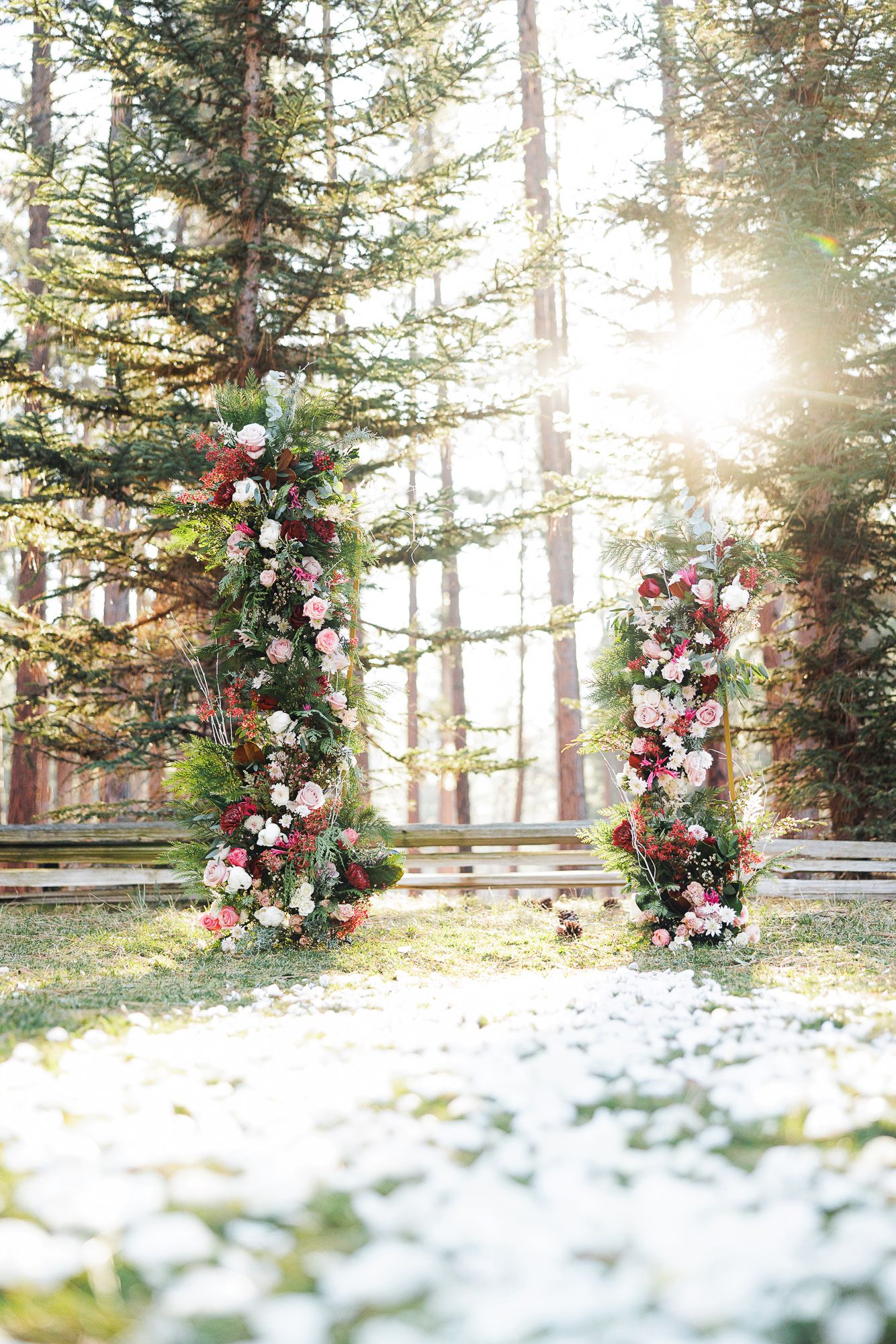Floral wedding arch with pink and white flowers stands in a snowy forest setting with evergreen trees and sunlight.