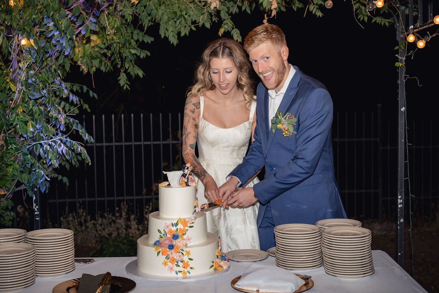 A couple cuts their floral-decorated wedding cake together during an outdoor evening reception.
