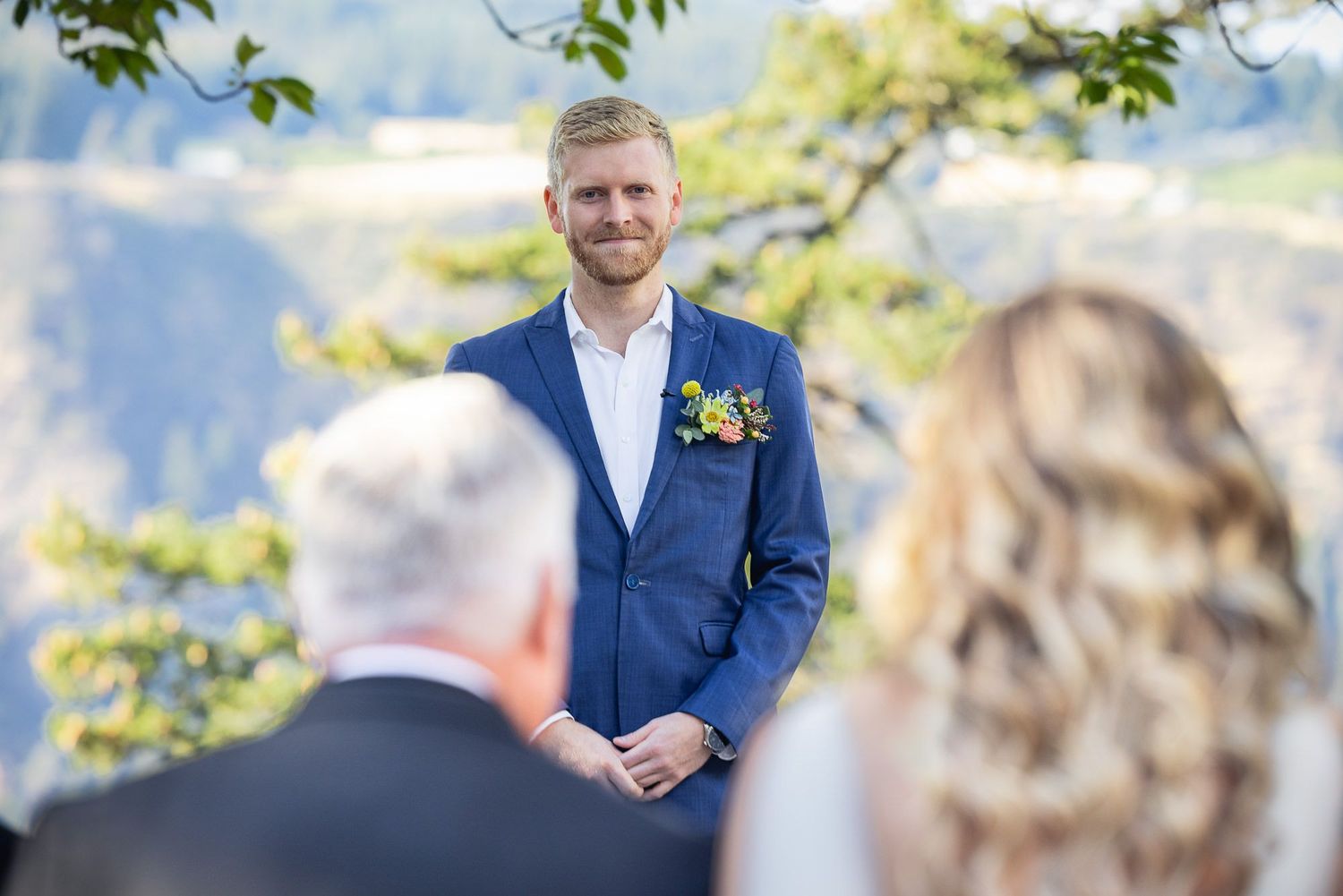 A groom in a navy suit with yellow boutonniere smiles during an outdoor wedding ceremony.