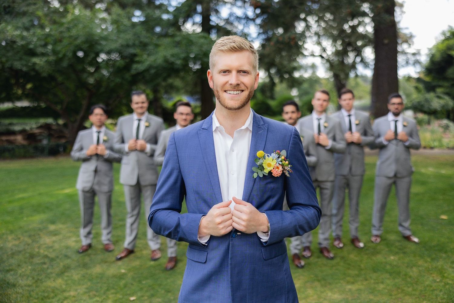 A groom in a navy blue suit with yellow boutonniere stands smiling while groomsmen pose in gray suits in the background.
