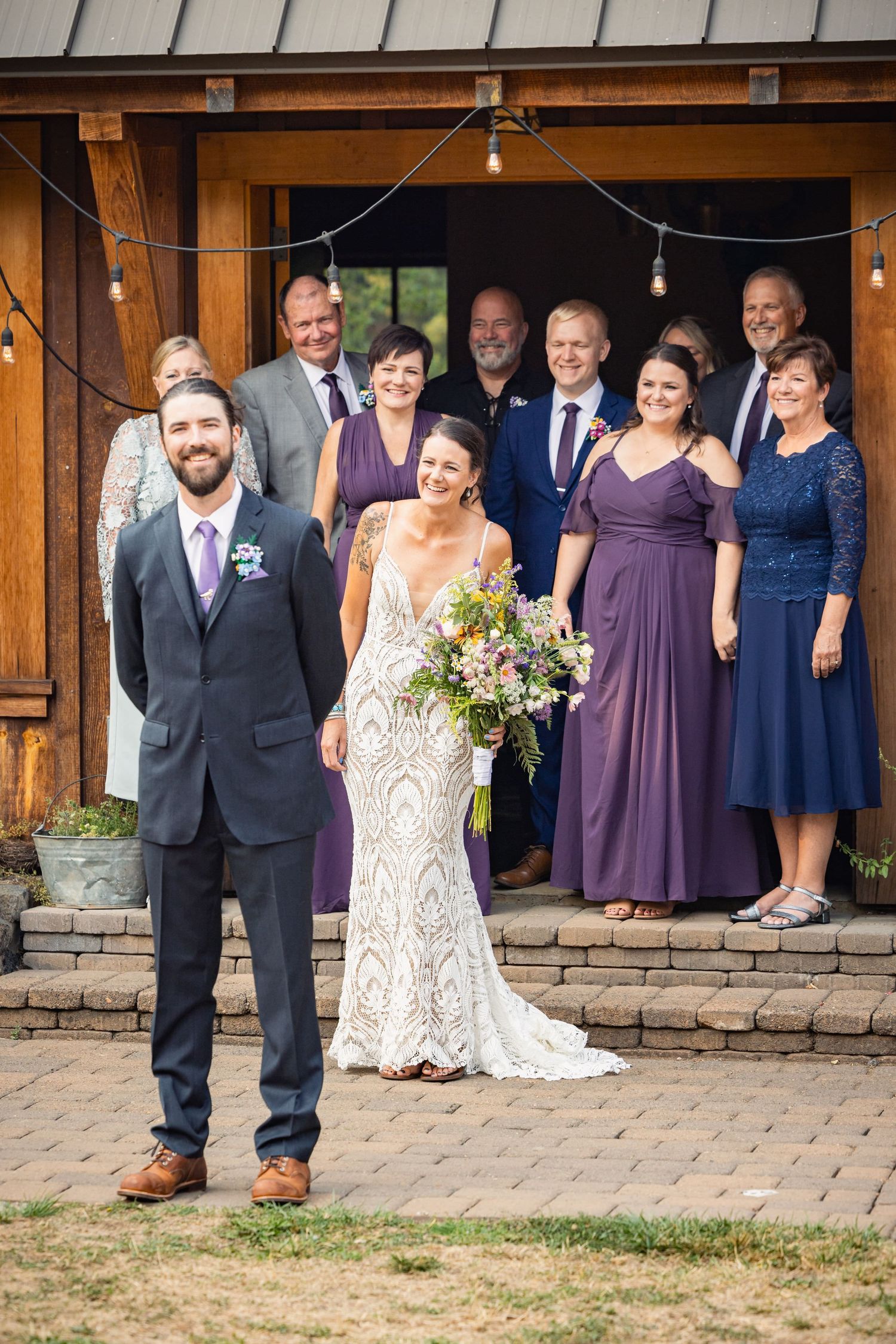 Wedding party poses in front of rustic wooden barn backdrop with string lights and stone foundation.