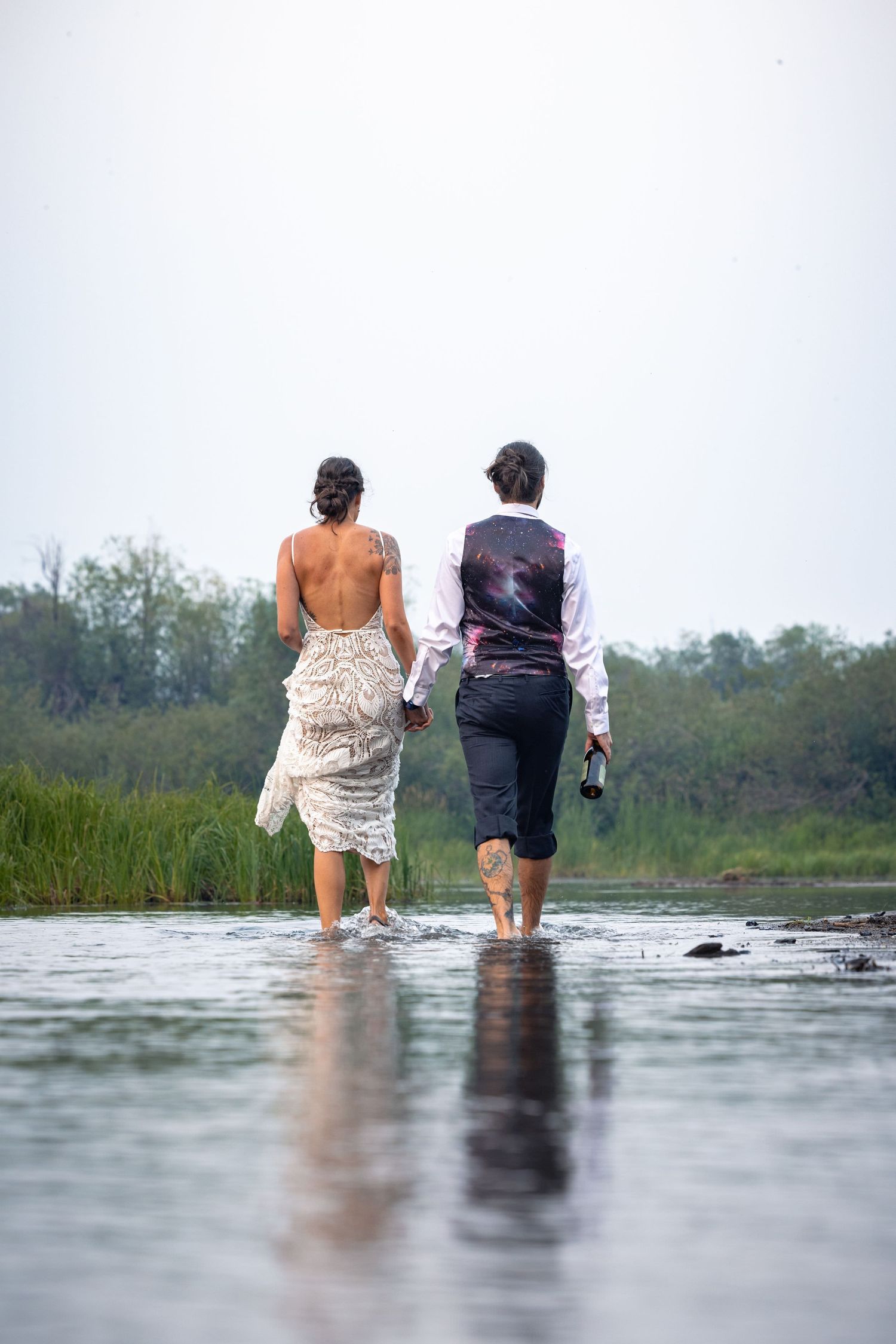 Two people in formal attire wade through shallow water together, creating reflections on the calm surface below.