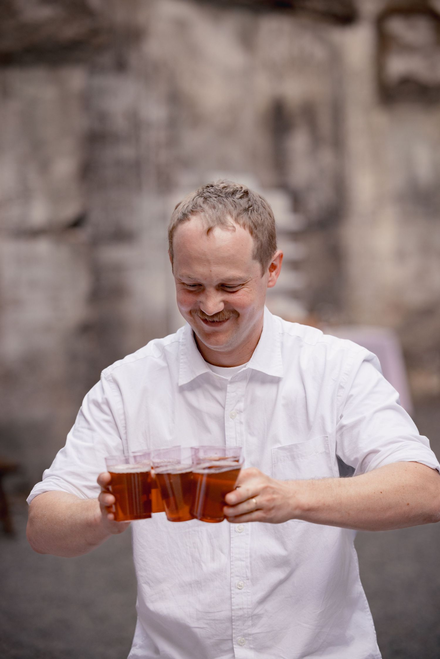 Person in white dress shirt smiling while carrying multiple plastic cups of beer outdoors.