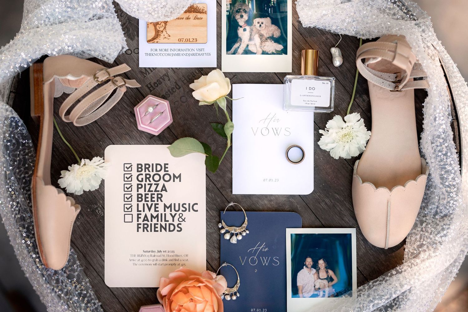 Flat lay of wedding details including blush shoes, invitation, photos, lace veil, and peach and white roses on dark background.
