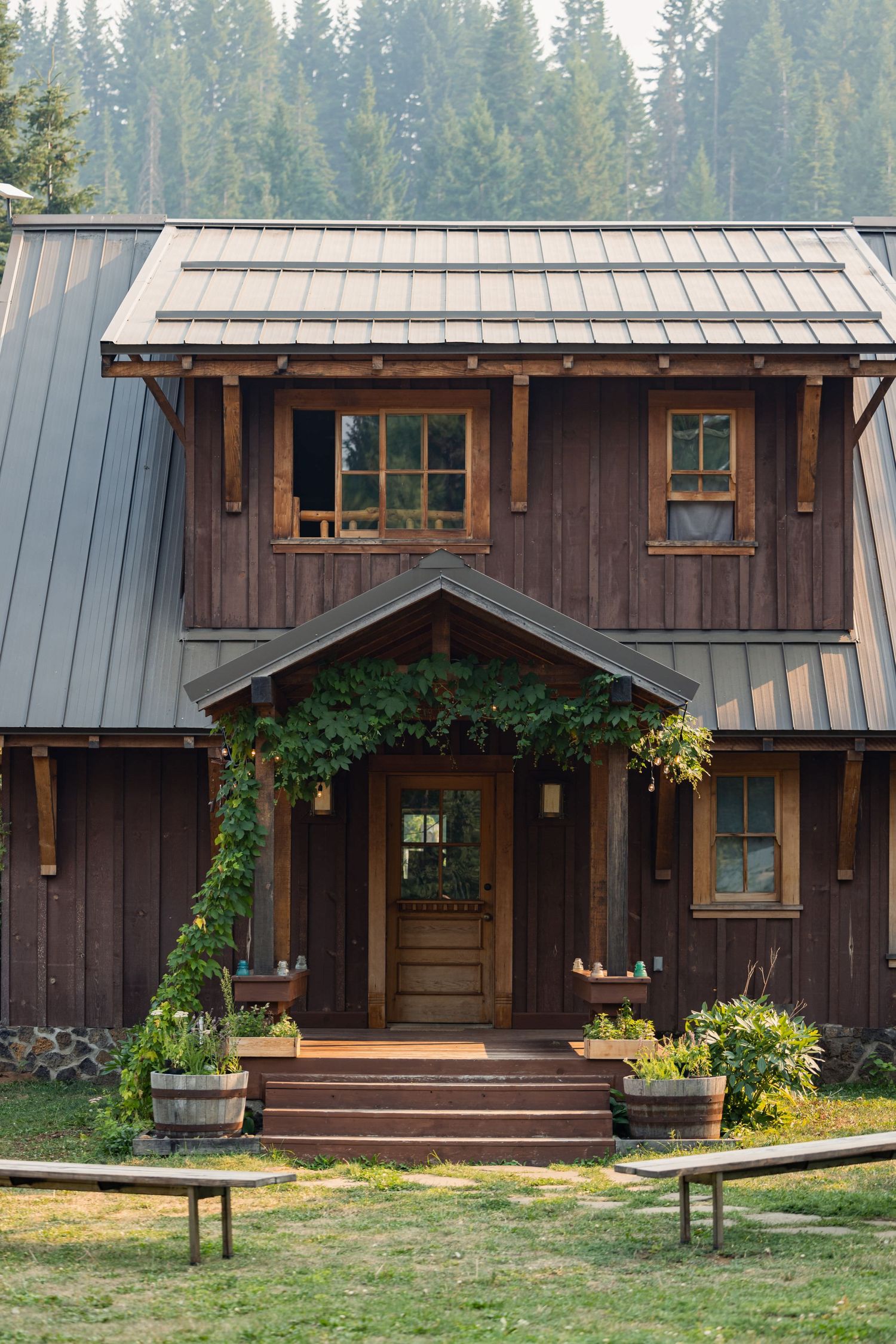 A rustic brown wooden cabin with metal roof features climbing vines, wooden windows, and potted plants on the front porch steps.