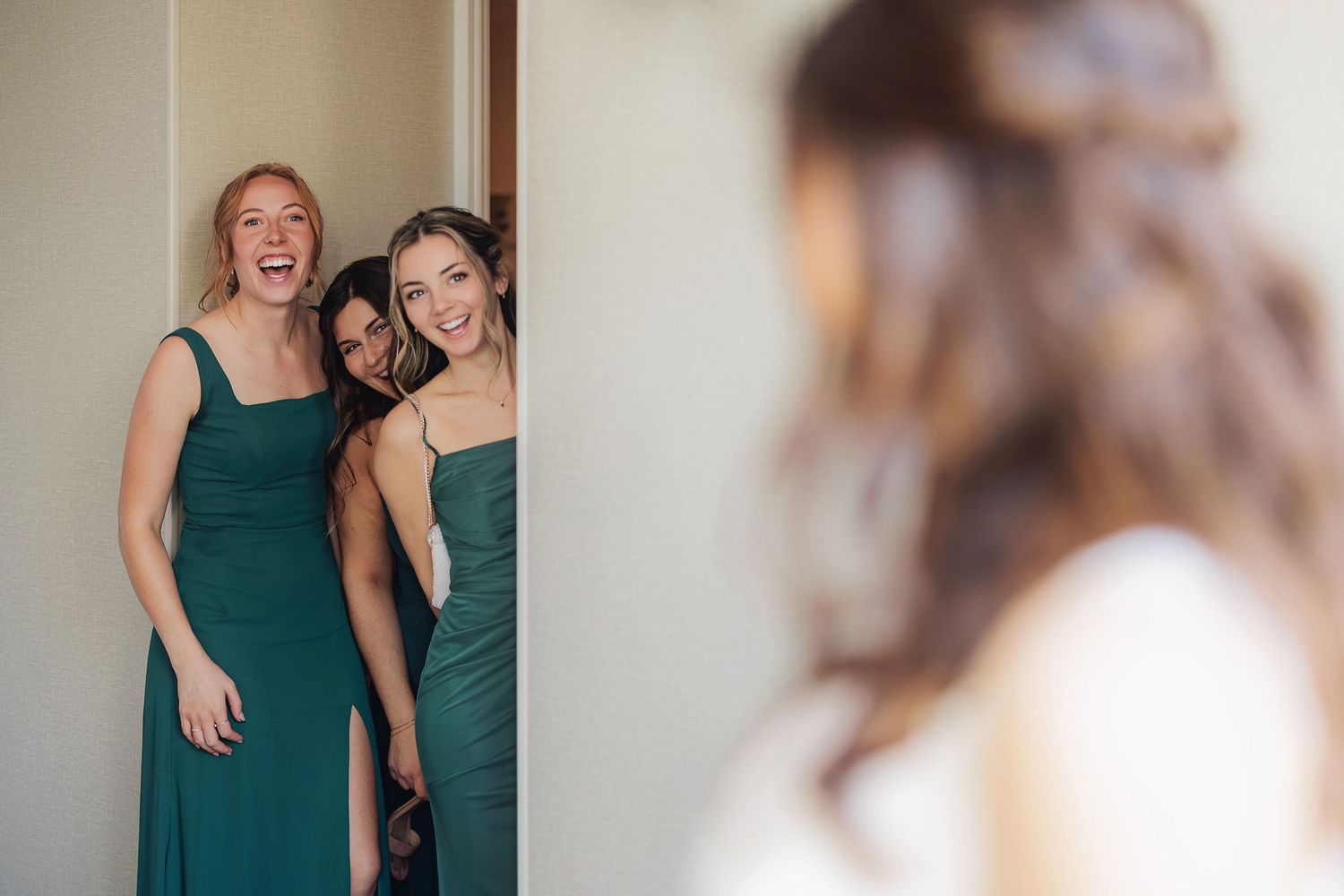 Bridesmaids in emerald green dresses share a joyful moment while peeking around a corner.