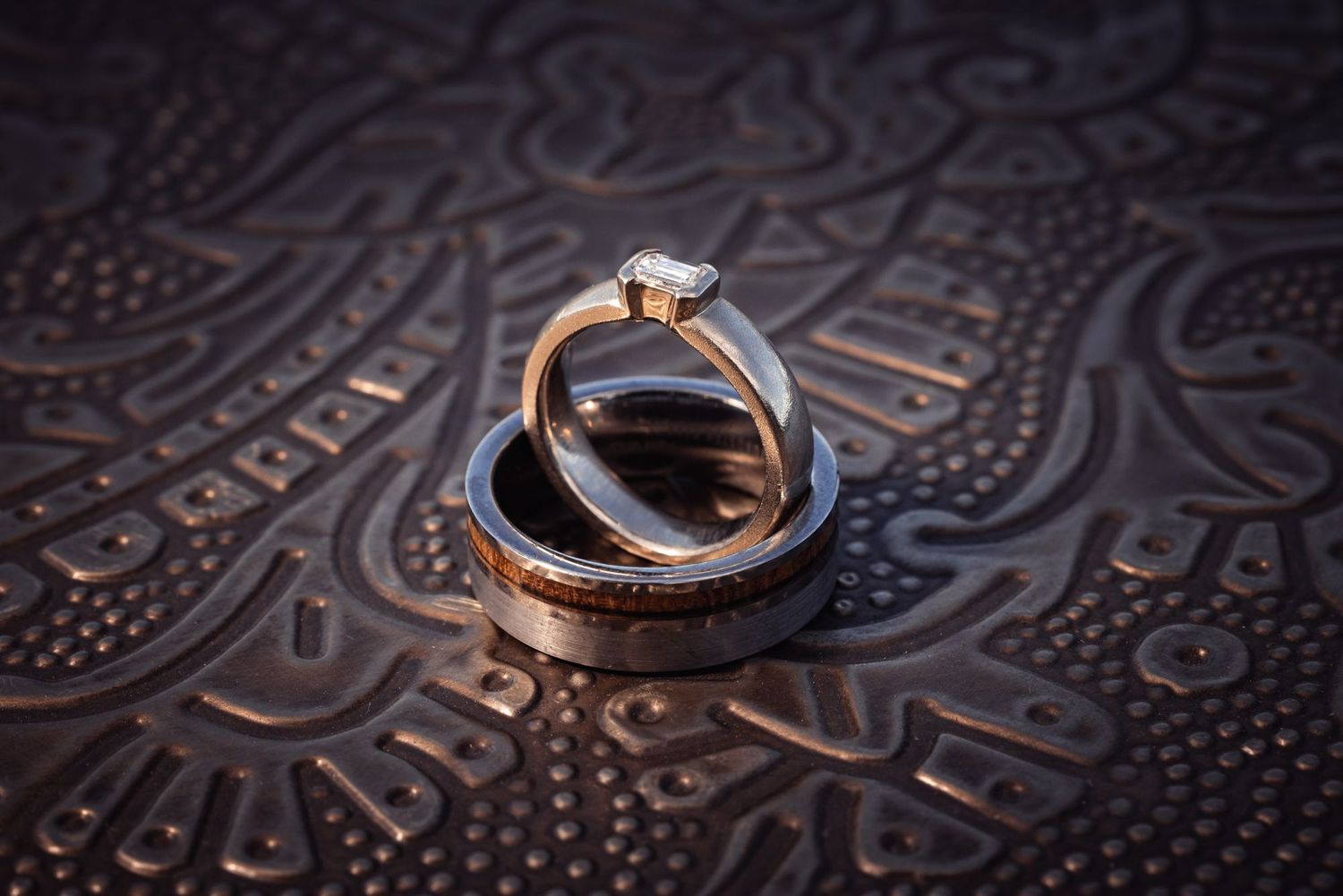 Two wedding bands and a diamond engagement ring rest on an ornate leather embossed surface with intricate patterns.