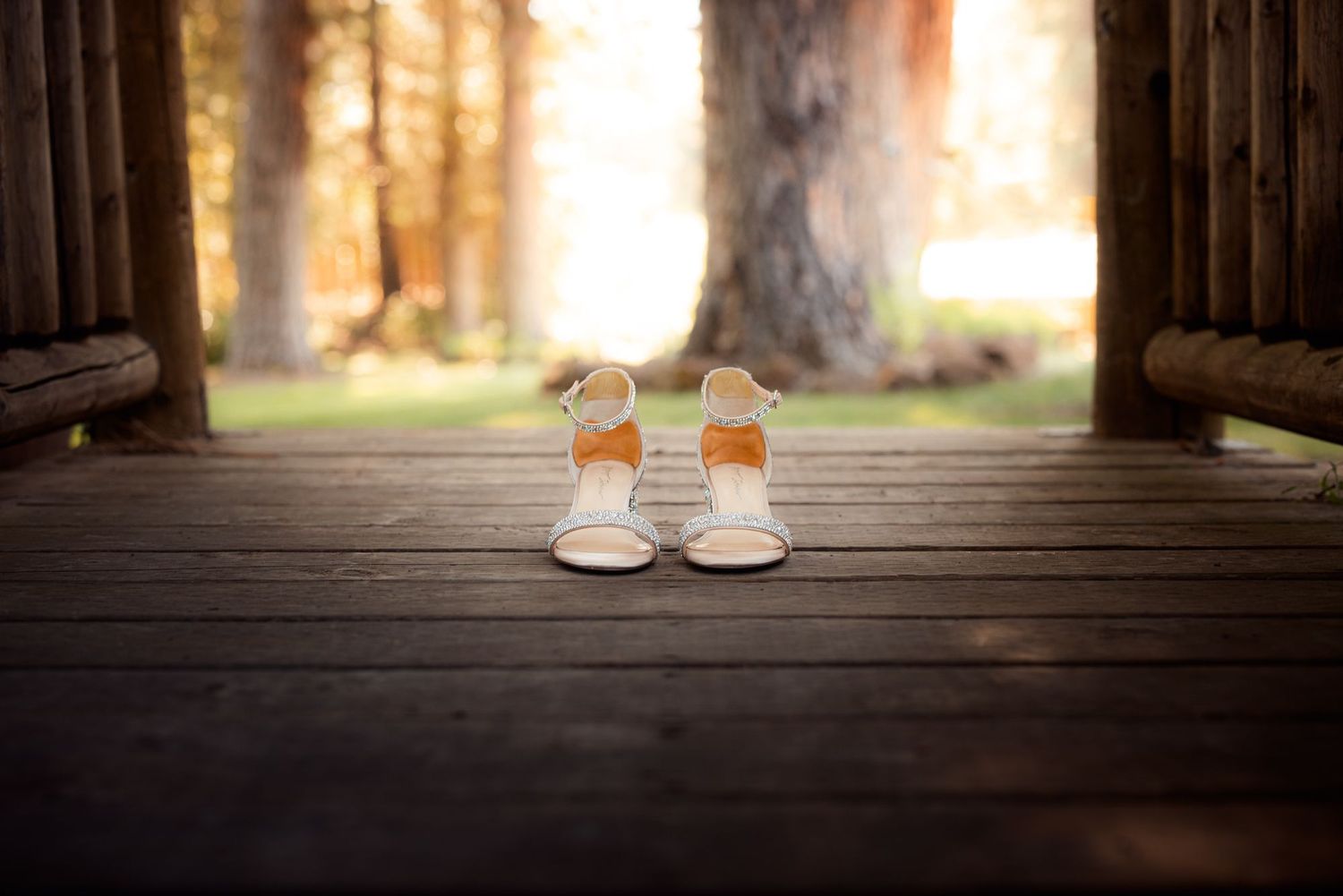 Small white baby shoes sit on a wooden deck with sunlit forest trees in the background.