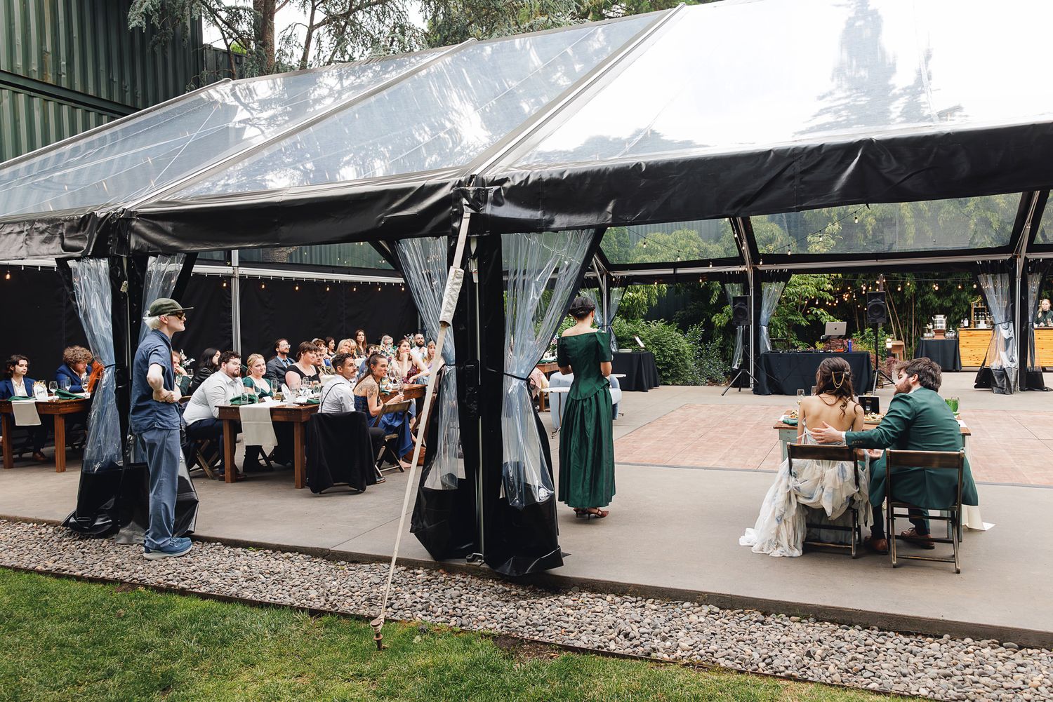 Outdoor wedding reception under a clear tent with guests dining at long tables, surrounded by lush greenery.