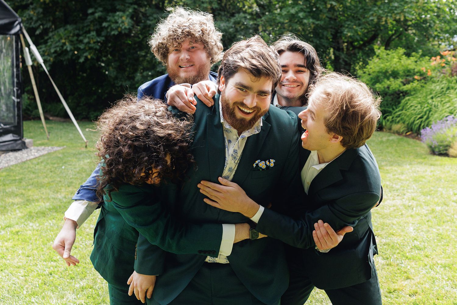 Group of friends in green suits laughing and embracing outdoors in a garden setting.