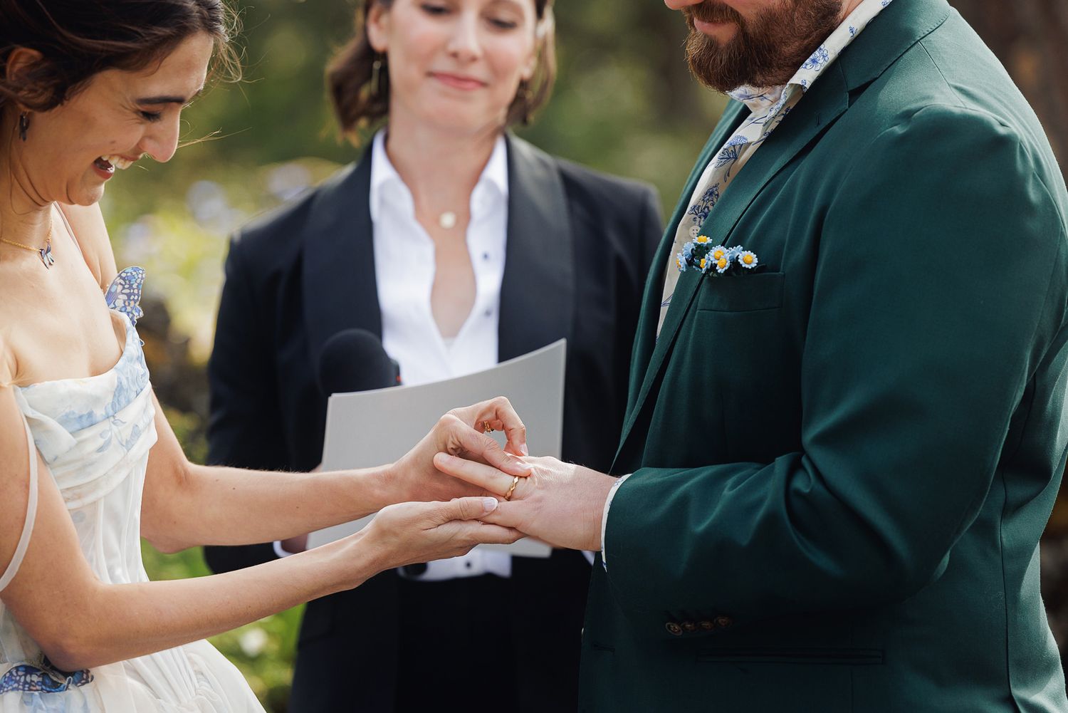 Bride placing wedding ring on groom's finger during outdoor ceremony, officiant holding script in background.