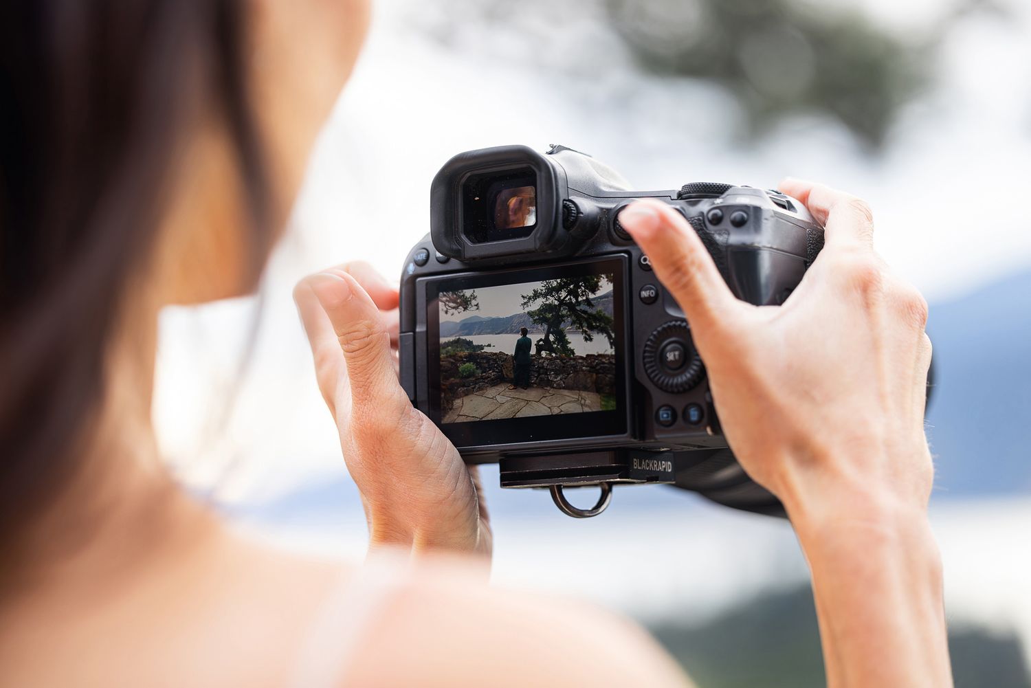 Woman holding a DSLR camera, photographing a scenic landscape displayed on the camera's LCD screen outdoors.