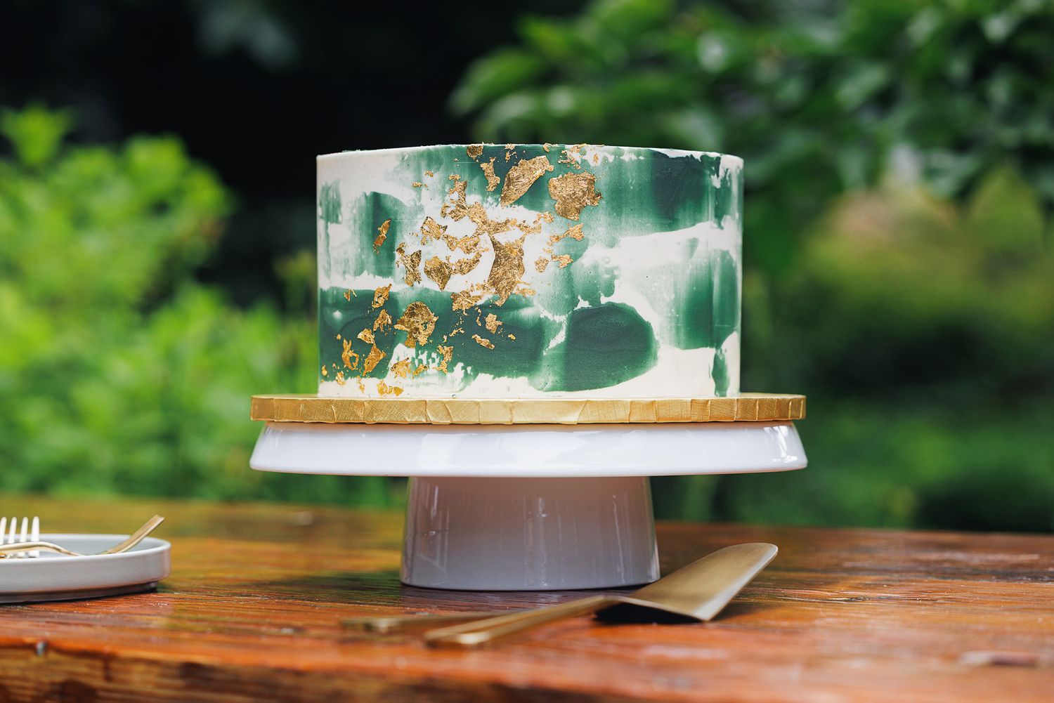 Elegant green and gold marble fondant cake on a wooden stand outdoors with lush greenery background.