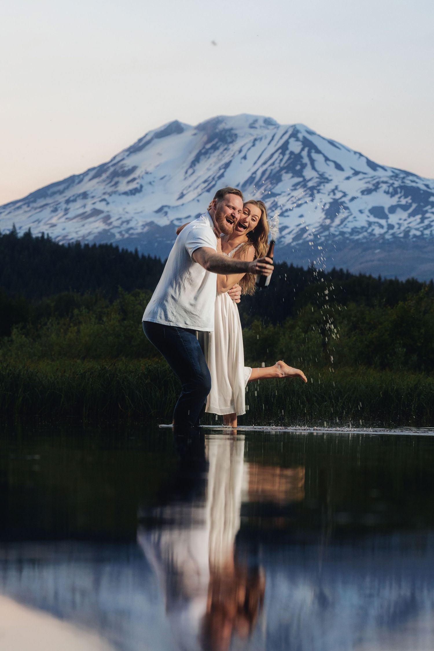 Couple laughing and splashing in a lake with snow-capped Mount St. Helens reflected in the water at sunset.
