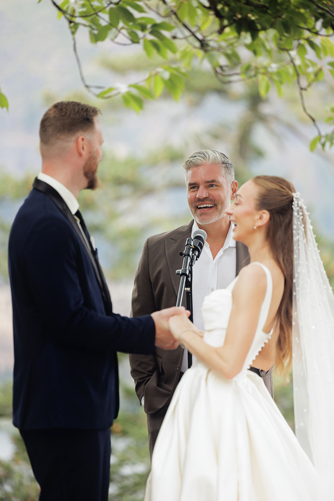 Bride and groom holding hands at outdoor wedding ceremony with smiling officiant, lush green trees in background.
