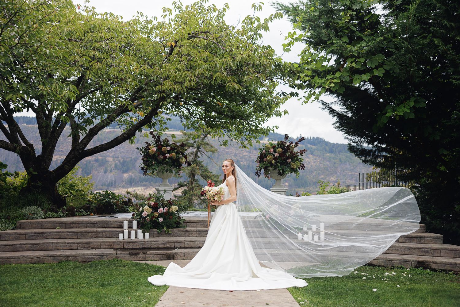 Bride in flowing white gown and veil poses in garden with floral arrangements and mountain backdrop.