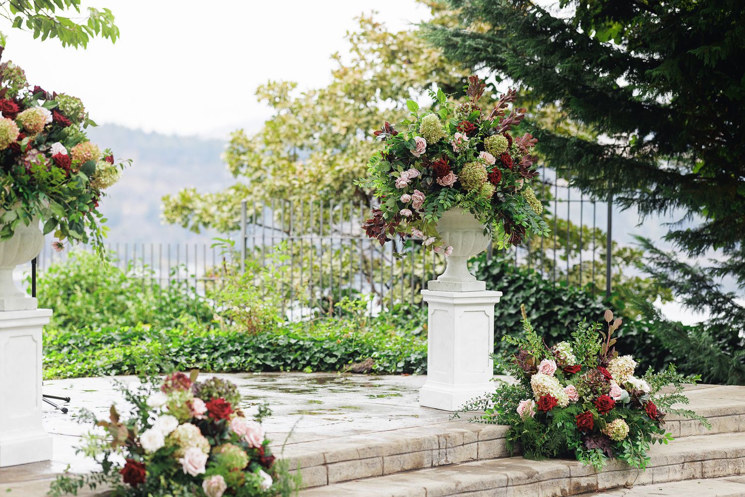 Elegant outdoor wedding arch adorned with lush floral arrangements in red, pink, and white blooms against a scenic garden backdrop.