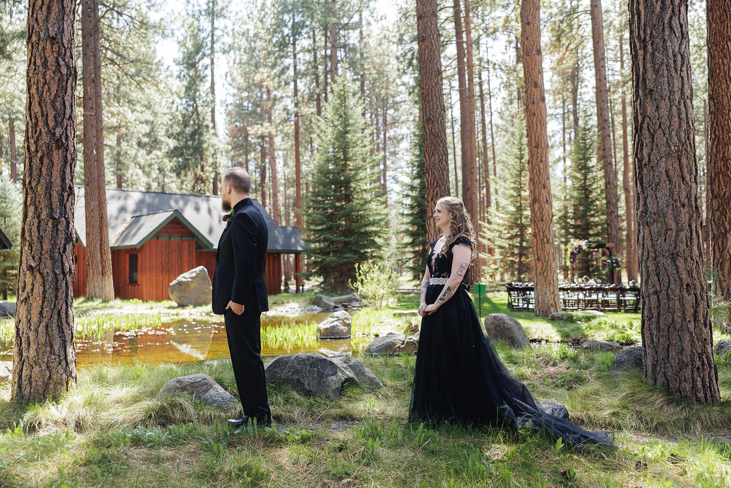 Couple in formal attire standing apart in a pine forest with a red cabin backdrop for an outdoor engagement session.