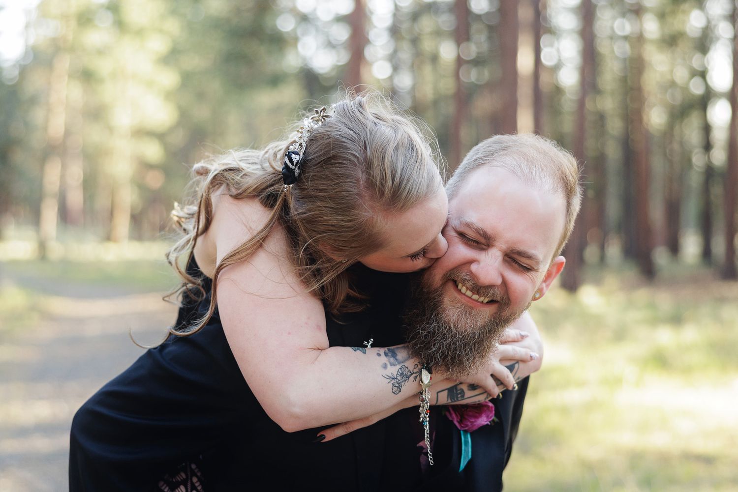 Bride hugging groom from behind on a forest path, both smiling joyfully during an outdoor wedding photo session.