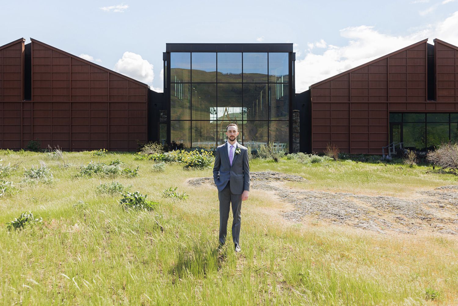 Man in suit stands before modern rustic building with wood cladding and glass facade surrounded by wild grass.