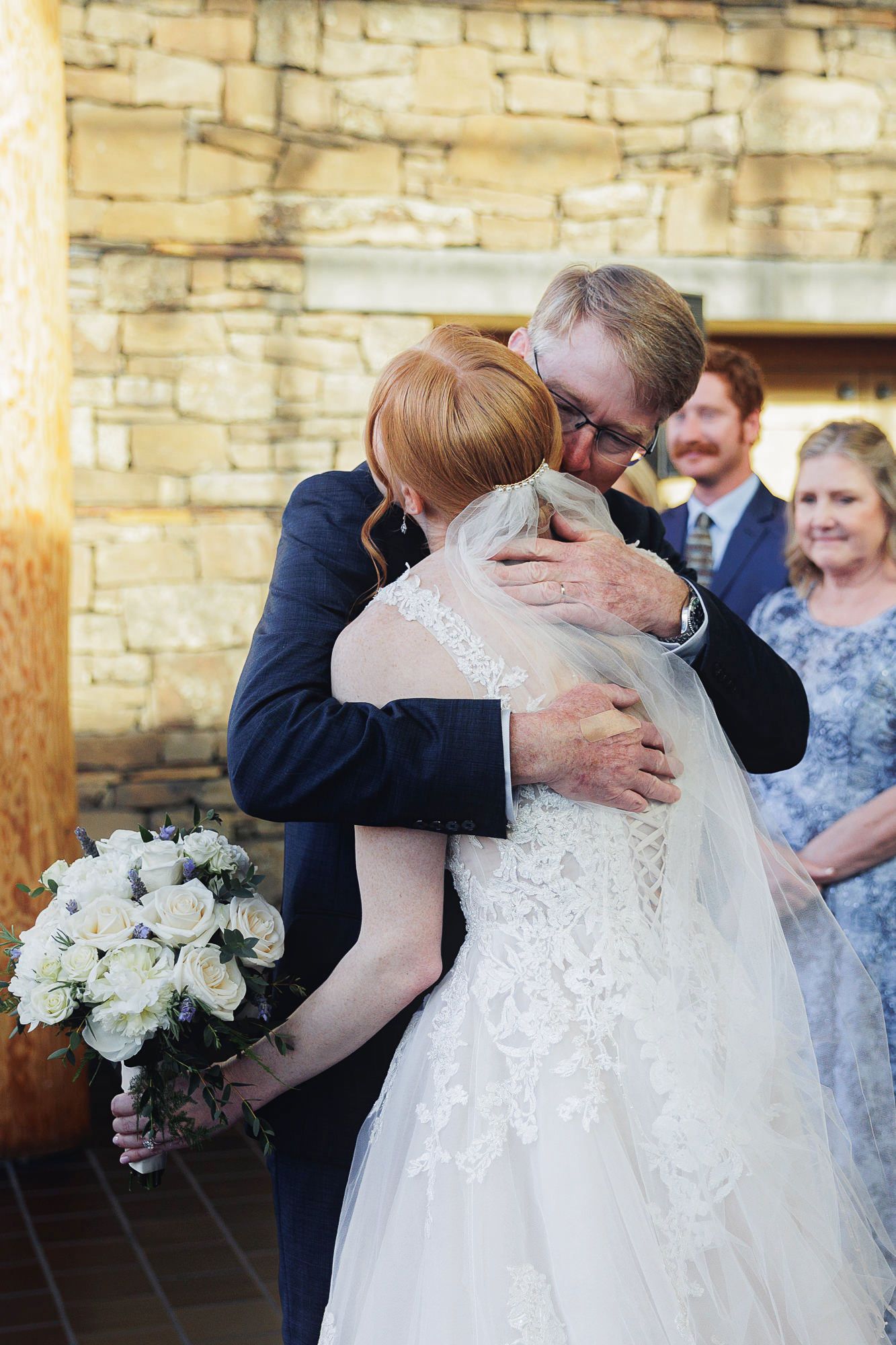 Bride and groom share emotional hug at wedding ceremony, bride holding white rose bouquet in lace gown.