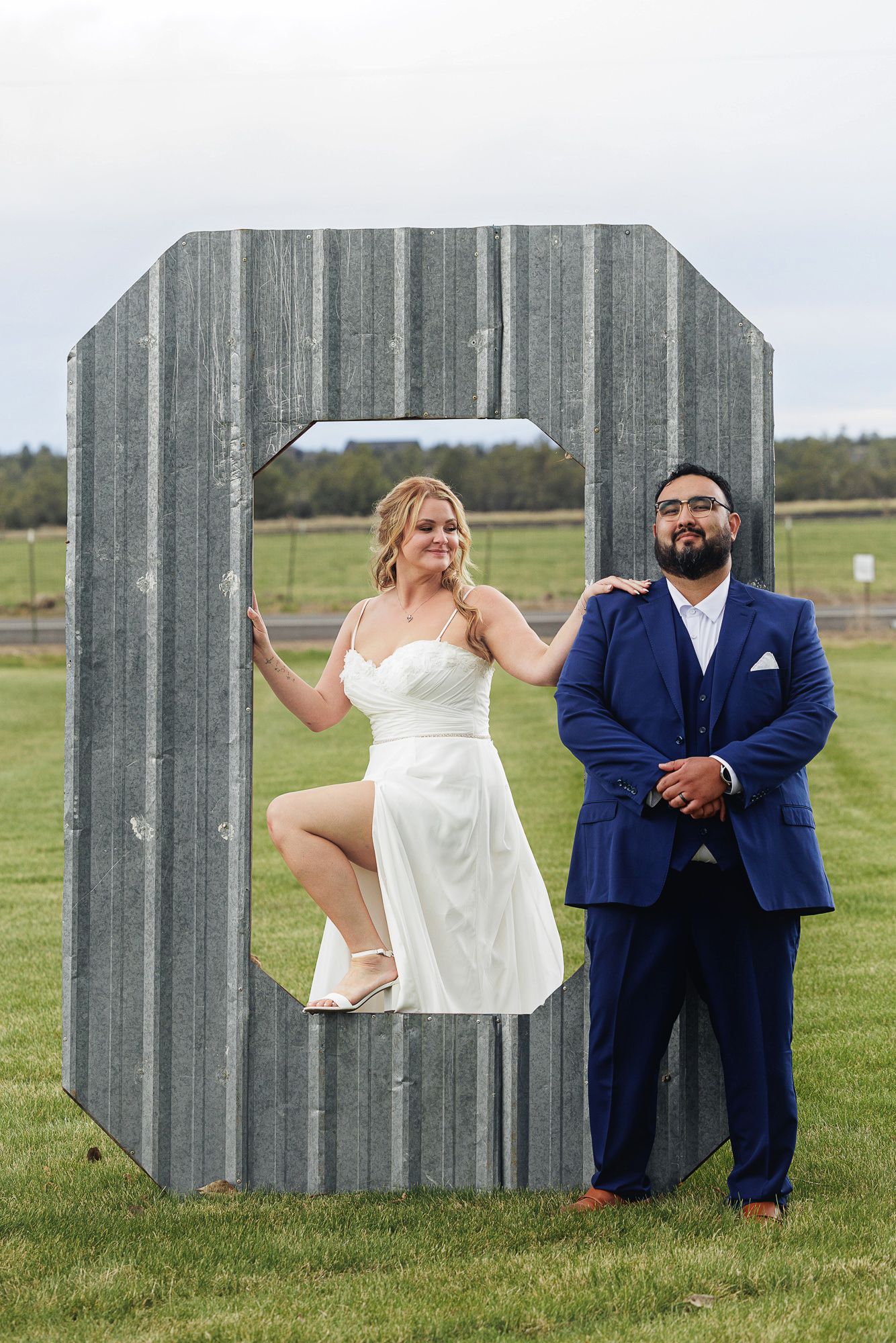 Bride and groom posing with a large metal letter C on a green lawn, wearing white dress and navy suit.