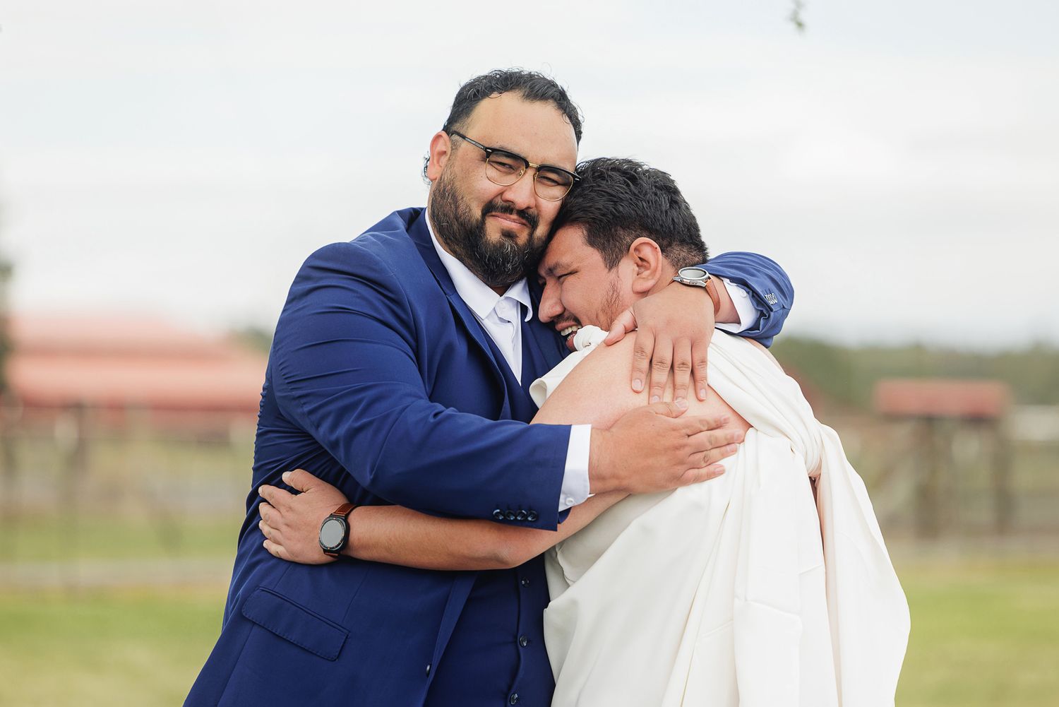 Two grooms share a warm embrace during their wedding ceremony outdoors at a rustic farm venue.