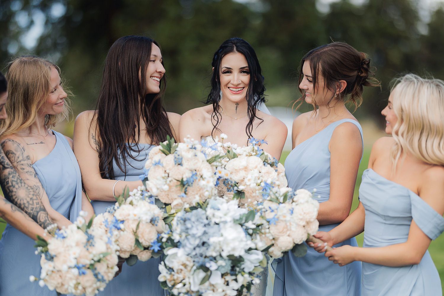 Bridesmaids in light blue dresses holding white and blush floral bouquets gather for photos during outdoor wedding.